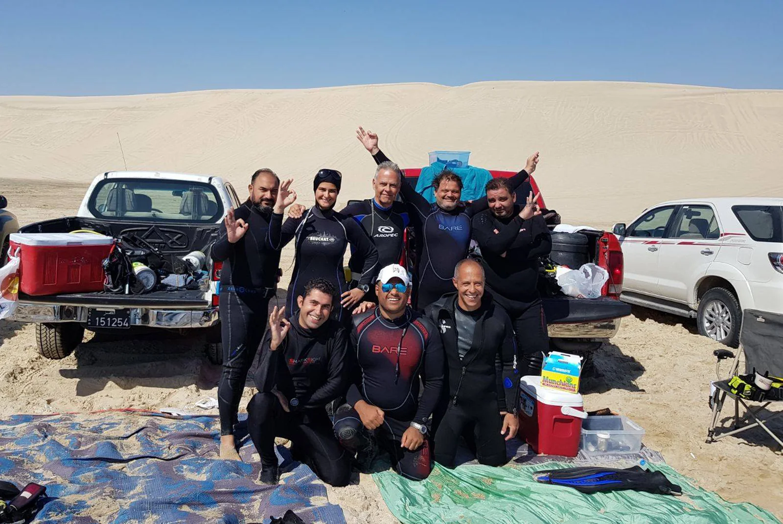 A diving team posing with their equipment beside coastal sand dunes