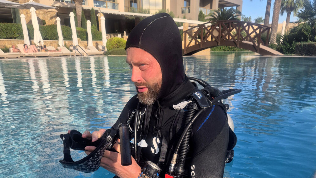 A PADI diving professional in full scuba equipment prepares his mask beside a swimming pool at a dive training facility. Wearing a black hooded wetsuit and BCD, he carefully inspects his mask while standing in the water. The sunlit surroundings, poolside loungers, palm trees, and resort-style buildings create a relaxed setting for professional dive training.